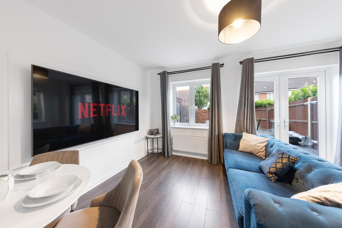 A modern living area features a large television displaying the Netflix logo. Comfortable seating is provided by a blue sectional sofa adorned with decorative pillows. Natural light enters through two windows, framed by elegant curtains, creating a cozy ambiance for relaxation.