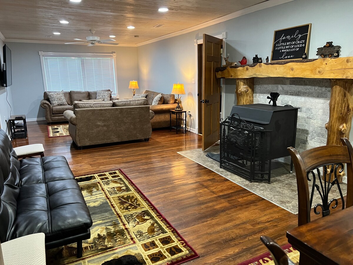 A welcoming living area features a mix of comfortable seating arrangements, including a sofa, chairs, and a futon. A rustic fireplace adorned with decorative elements serves as a focal point. Warm lighting is provided by lamps, enhancing the inviting atmosphere of the room.