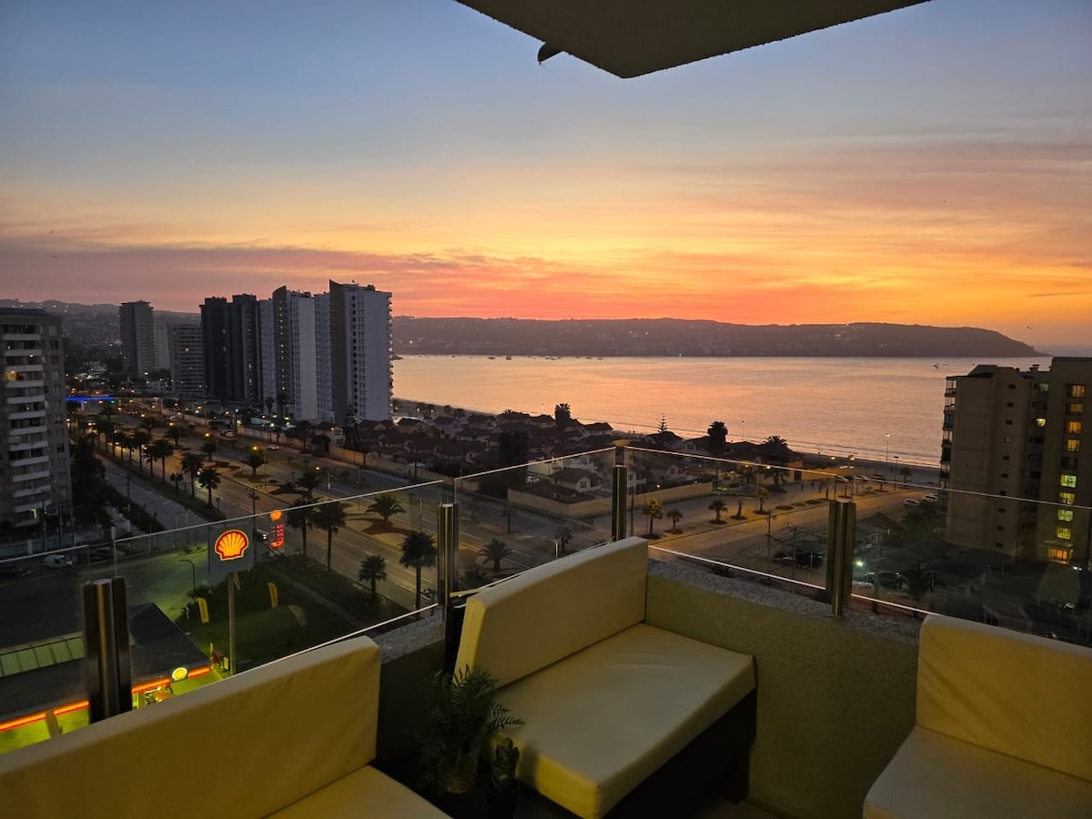A vibrant sunset is captured over the ocean, casting warm hues across the sky. In the foreground, a seating area features light-colored sofas, with a view of the coast and cityscape below. The scene showcases a tranquil ambiance, ideal for relaxation.