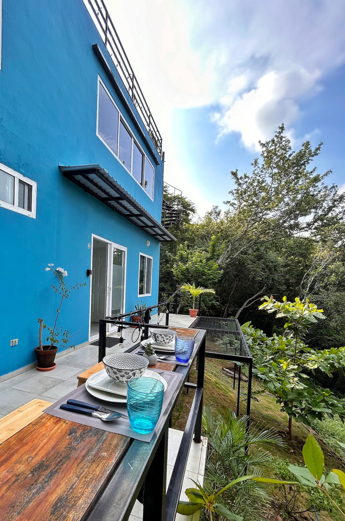 An outdoor dining area features a wooden table set with plates and stacked bowls, accompanied by turquoise glasses. The vibrant blue exterior of the building contrasts with the surrounding greenery and trees, creating a serene atmosphere. A glass railing provides a clear view of the natural landscape.