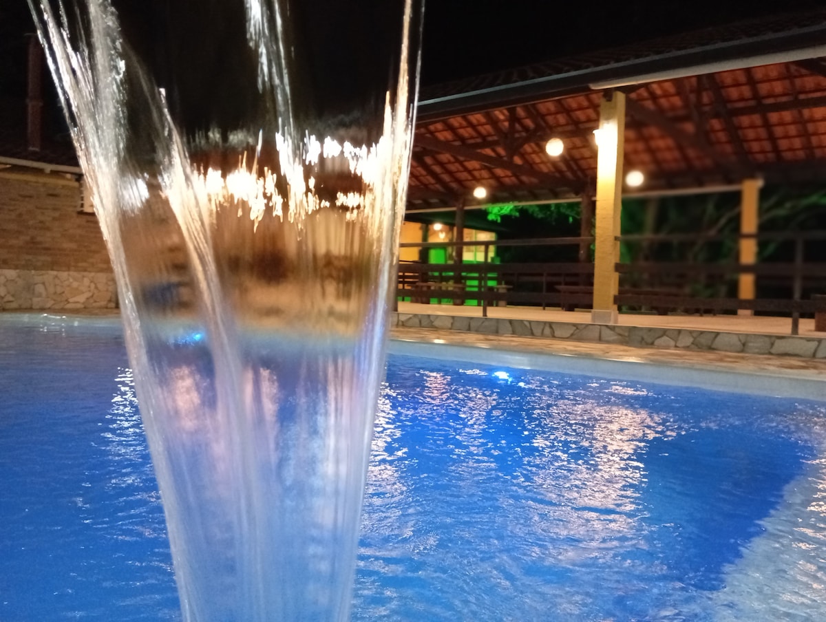 A serene swimming pool is illuminated under soft lighting, creating a calm ambiance. Water cascades gently from a fountain, creating ripples on the surface. A covered patio with warm wooden beams is visible in the background, reflecting a peaceful outdoor setting.