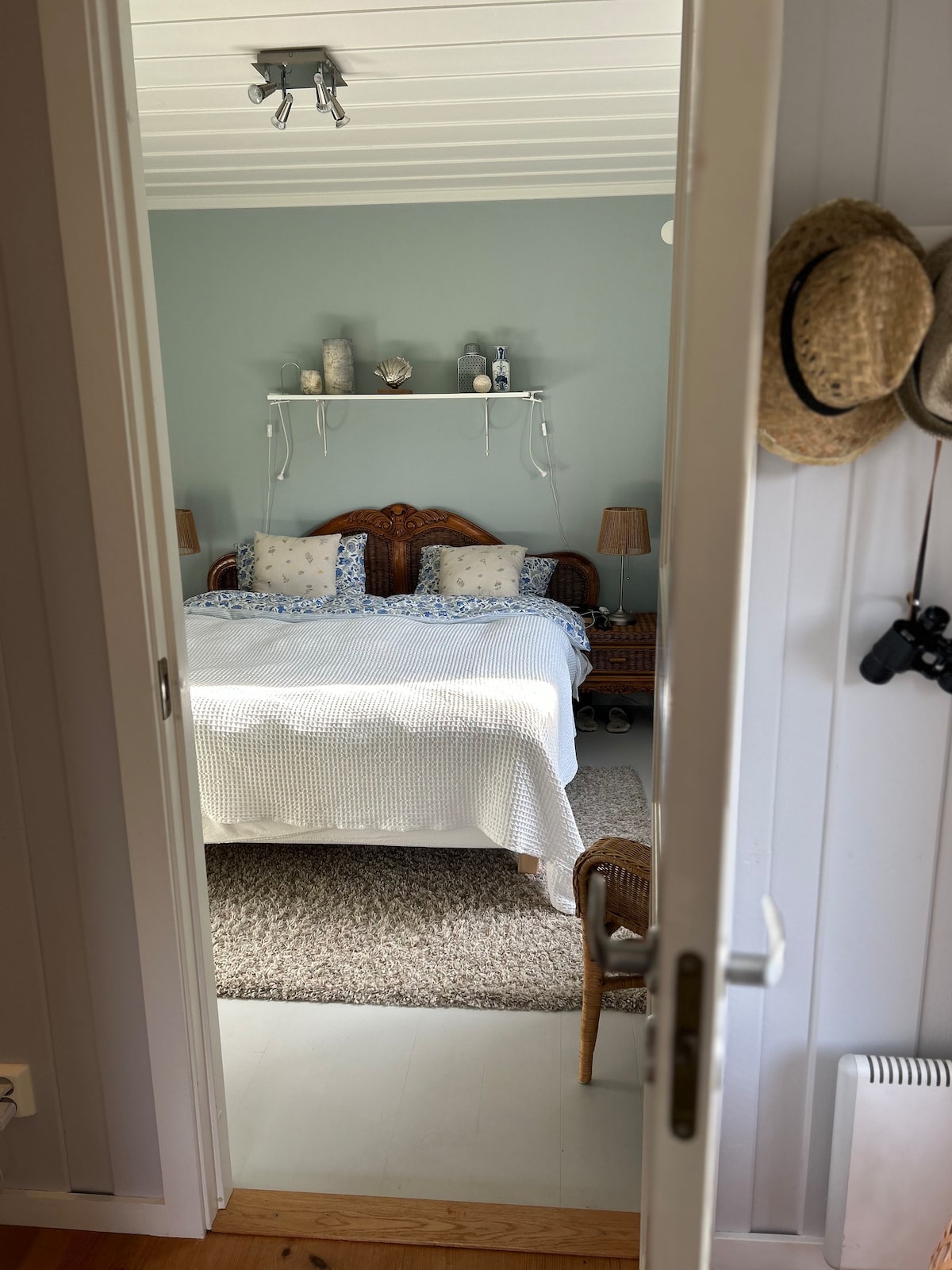 A spacious bedroom is visible through an open door, featuring a large bed dressed in a textured blanket. Two bedside lamps provide soft lighting, while decorative items are arranged on a shelf above the bed. A comfortable chair and a rug complement the inviting atmosphere.