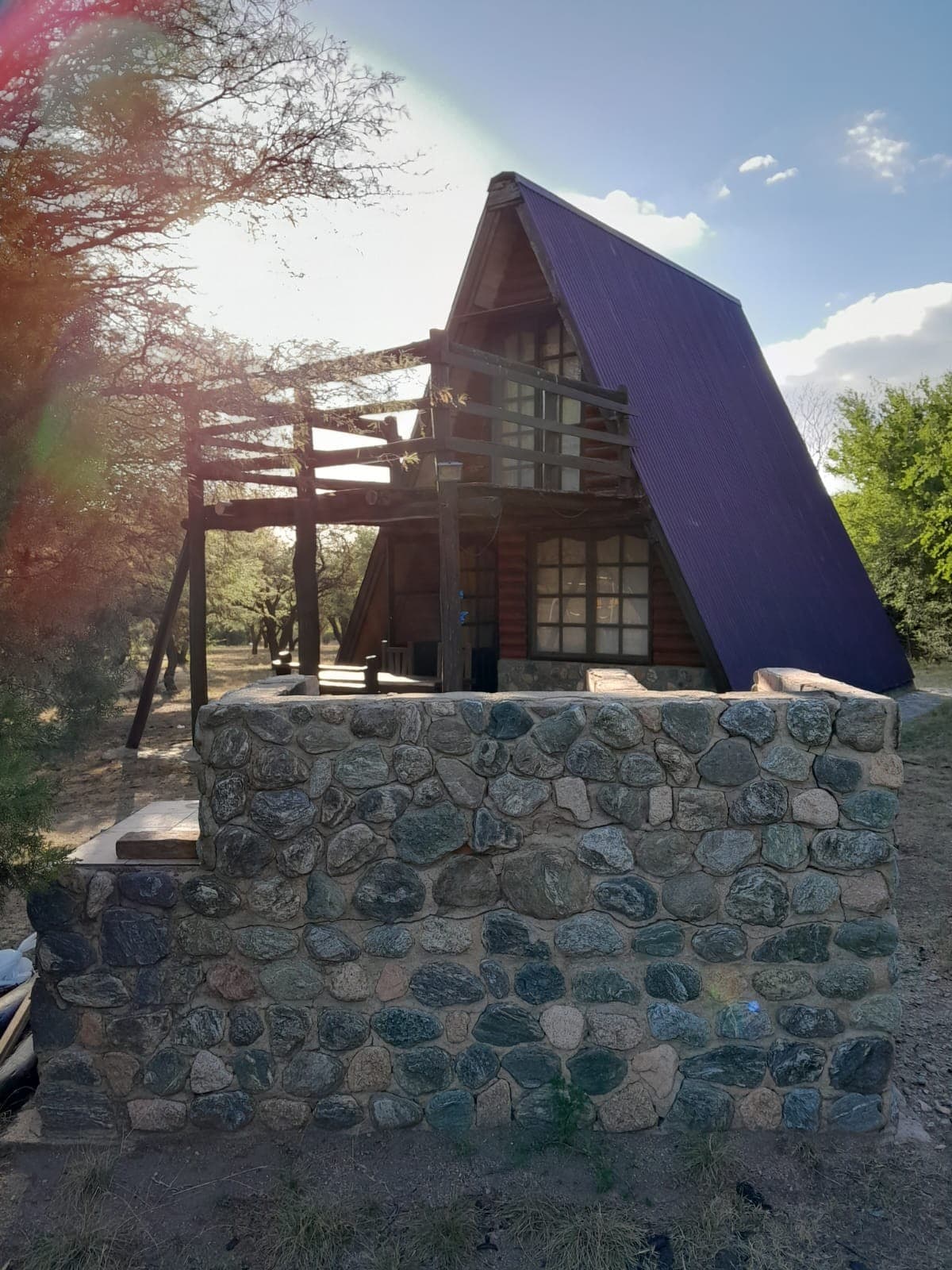 A charming A-frame cabin is nestled among trees, showcasing a unique purple roof. A stone wall surrounds the property, adding a rustic touch. Natural light filters through the trees, highlighting the cabin's wooden structure and inviting outdoor space.