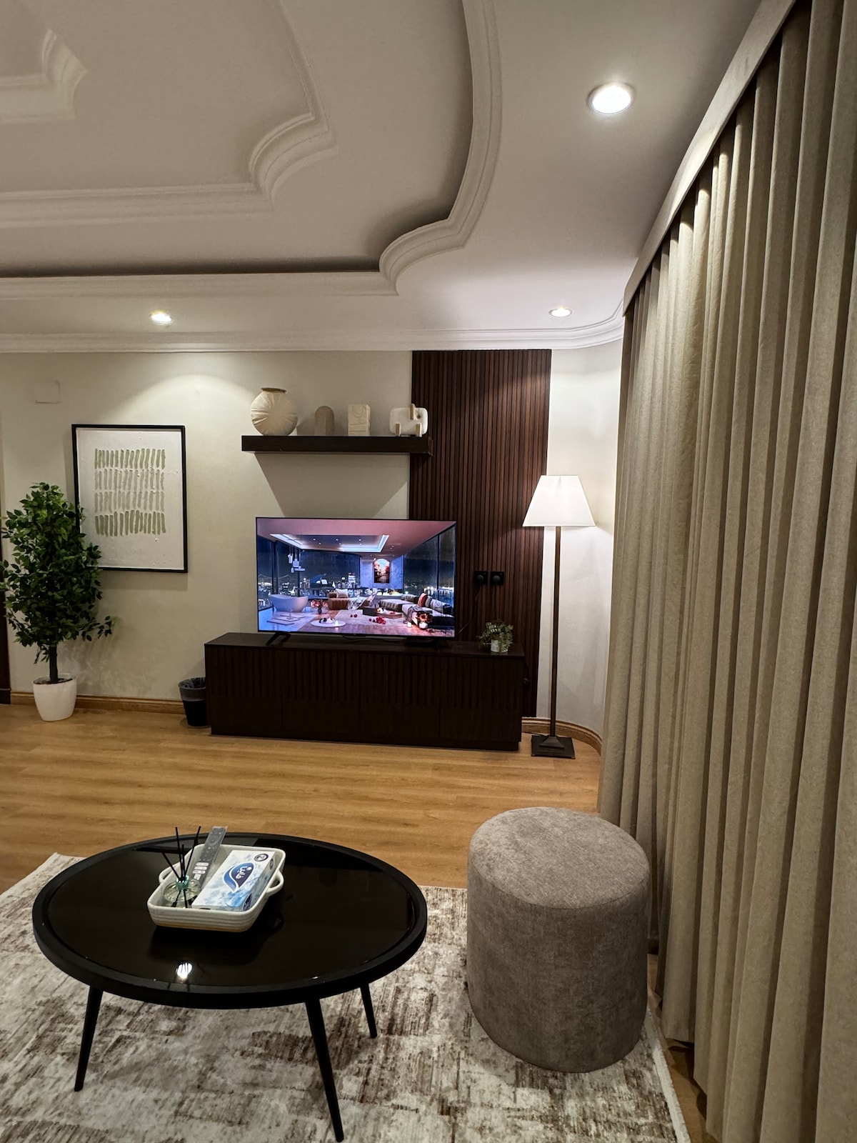 A comfortable living area features a sleek entertainment center with a modern smart TV. A round coffee table sits on a textured rug, accompanied by a plush ottoman. Natural light is filtered through elegant curtains, enhancing the warm tones of the space.