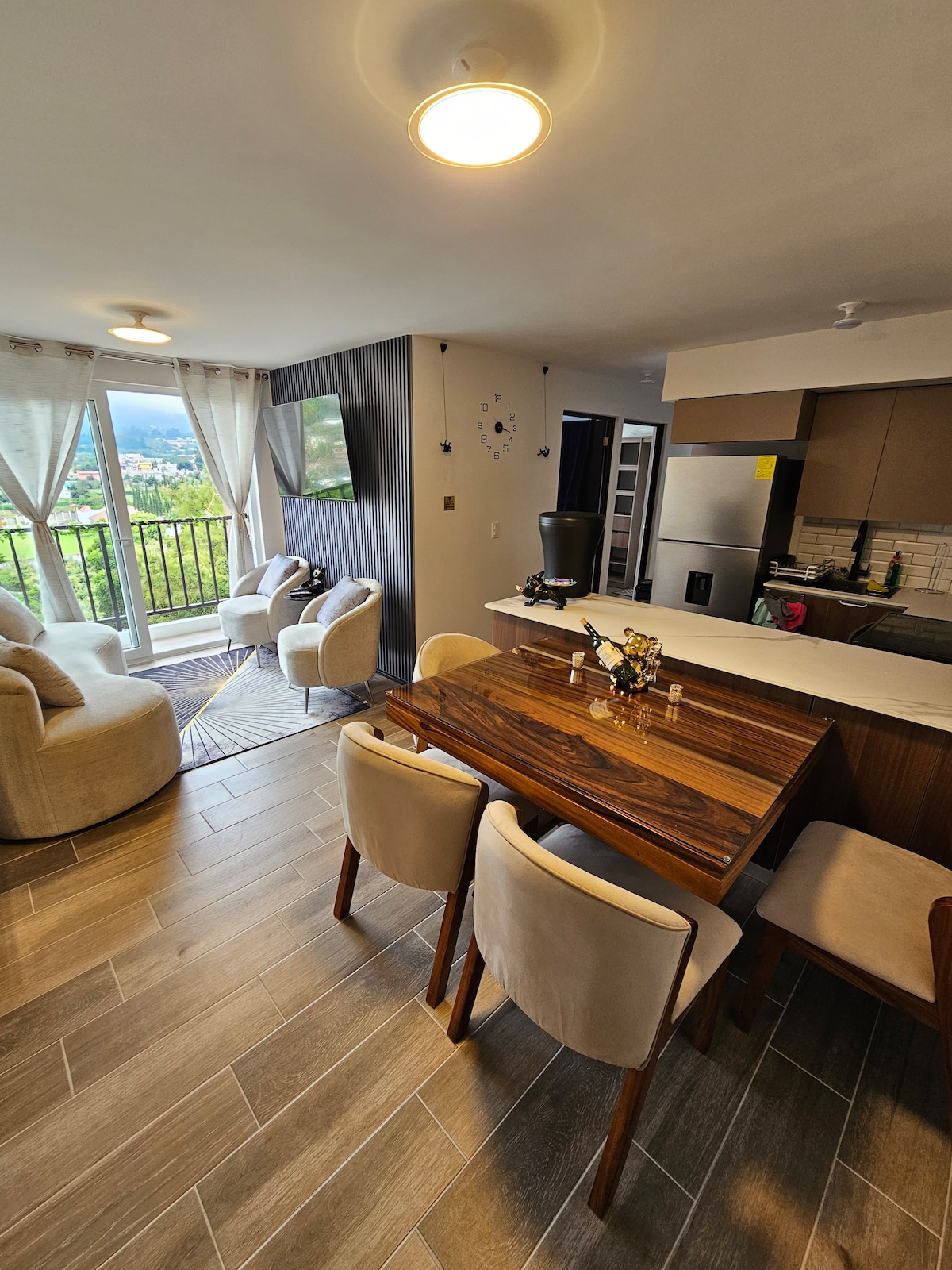 A modern dining area is featured with a wooden table surrounded by upholstered chairs. Soft, neutral-toned armchairs are positioned nearby. Natural light enters through large windows, illuminating the open-concept space, which includes a kitchen and living area, creating a cozy atmosphere.