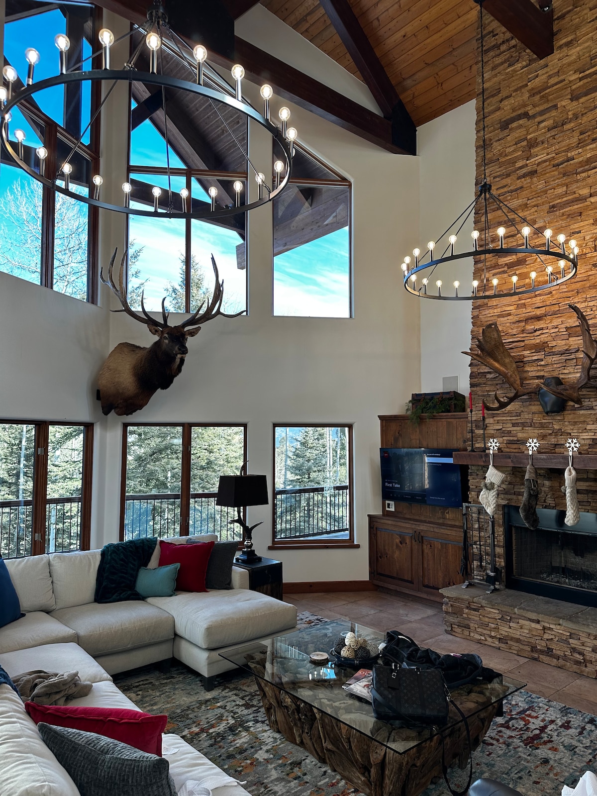 A spacious living area features high ceilings and large windows that provide views of the surrounding trees. A striking deer head adorns one wall, while two circular chandeliers hang above. A sectional sofa with colorful cushions surrounds a coffee table, creating a cozy gathering space.