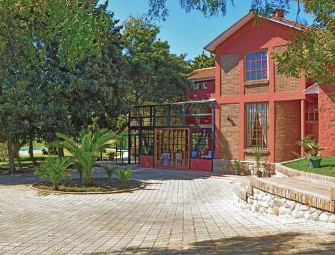 A charming exterior of the property is framed by lush greenery and trees. The red-brick building features large windows and a glass enclosure. The entrance is paved with stones, leading to a spacious outdoor area that includes landscaped gardens.
