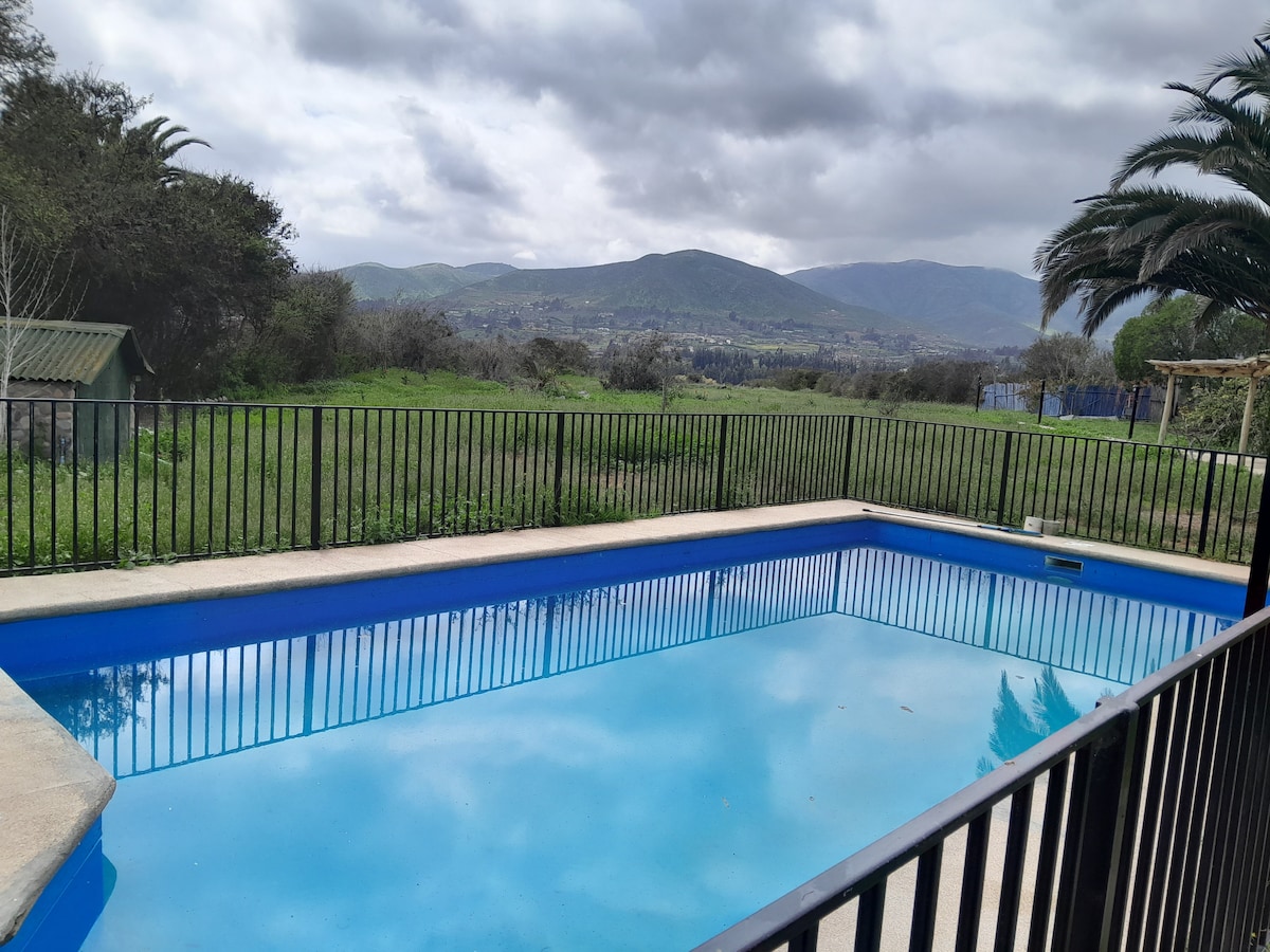 A clear blue pool is framed by a secure black railing, offering views of rolling hills and greenery. The cloudy sky adds a tranquil backdrop to the serene outdoor space.