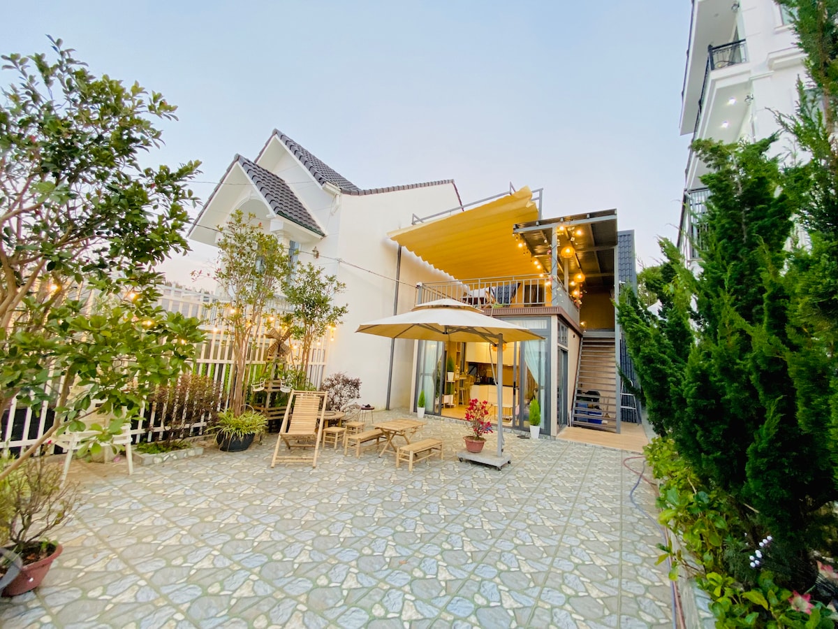 An outdoor terrace is highlighted by a spacious canopy, providing shade over wooden lounge chairs. The garden features various plants and flowers, enhancing the natural setting. A staircase leads to the upper level, while the exterior reflects a modern architectural style.