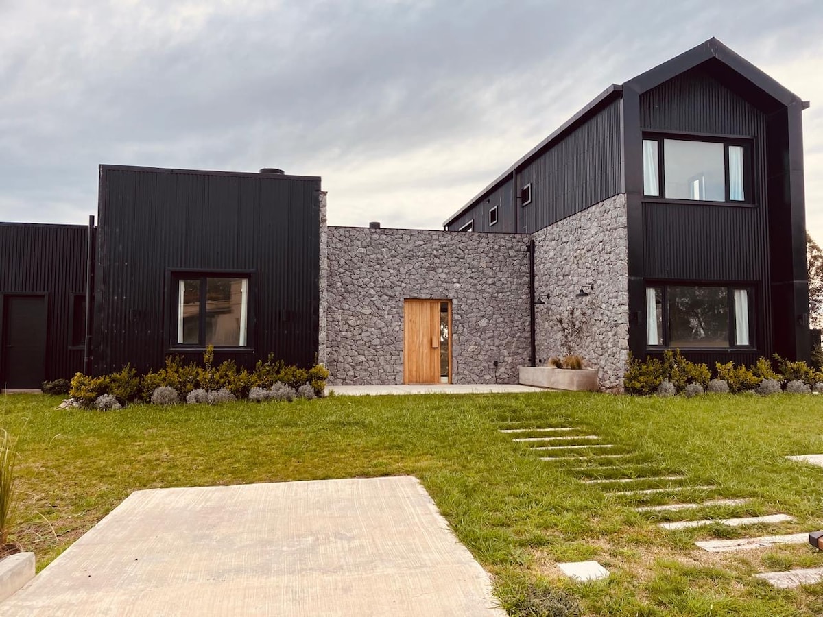 The modern exterior of the house features a combination of dark materials and natural stone. A wooden front door is framed by large windows, providing a contemporary look. The well-maintained lawn is accentuated by neatly placed stepping stones leading to the entrance.