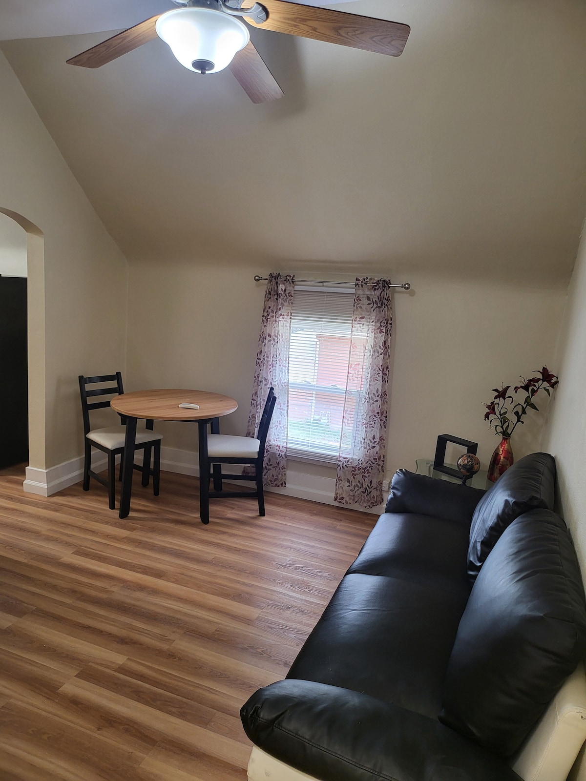 A cozy seating area is highlighted by a dark leather sofa. A round table with two black chairs occupies the space. Natural light enters through a window adorned with floral curtains, illuminating the warm wooden floor.