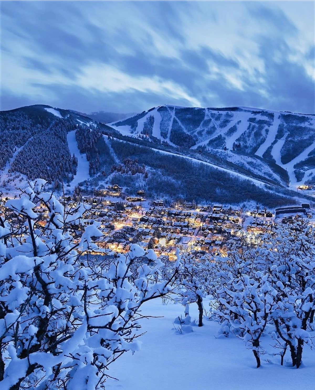 A winter landscape is captured, showcasing a snow-covered town nestled in a valley. Softly lit buildings glimmer among the snowy trees, with mountains rising in the background. Trails on the slopes are clearly visible against the serene backdrop of twilight skies.