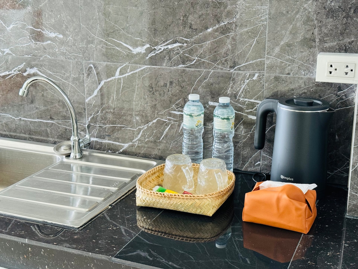 A kitchen area is shown with a stainless steel sink and a sleek dark countertop. Two bottles of water sit beside a kettle, with a woven tray containing two clear glasses and a small orange bag. The elegant marble wall adds a contemporary touch.