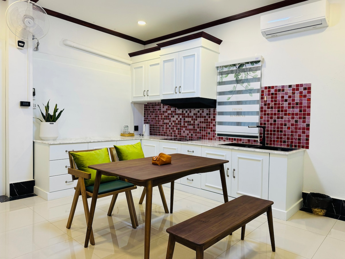 A modern kitchen area features white cabinetry with a red tiled backsplash. A wooden table with matching benches is positioned in the center, accompanied by green cushions. A small plant is placed on the countertop, with a ceiling fan and air conditioning unit visible.