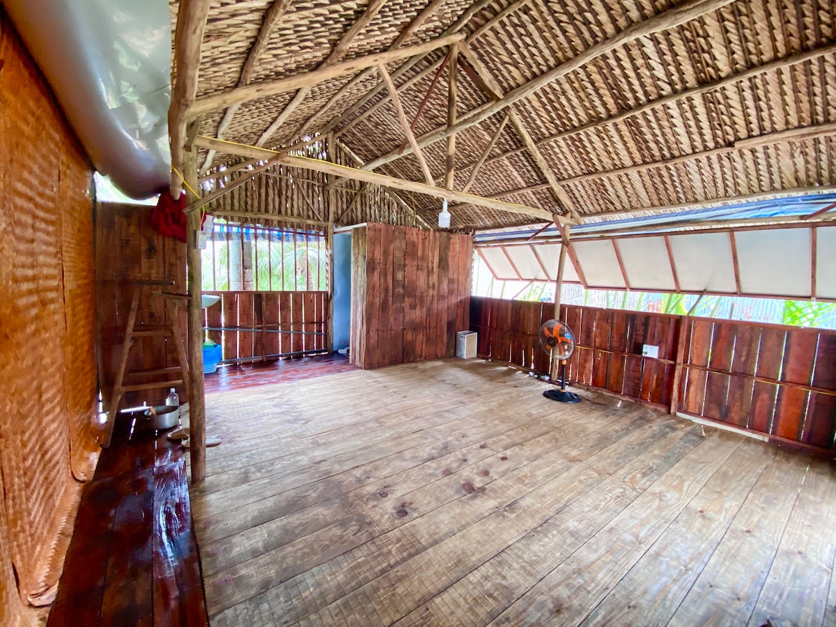 The interior space features a high, thatched roof supported by wooden beams. Natural light filters in through large windows, illuminating the polished wooden floor. The walls are constructed from wooden panels, creating a warm environment. A fan is positioned in one corner to provide airflow.