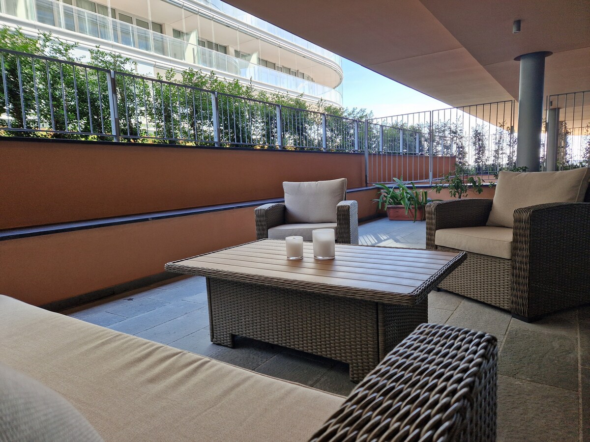 A covered outdoor seating area features a table and comfortable chairs made of woven materials. Lush greenery lines the space, providing a sense of privacy. The area is designed for relaxation, with natural light filtering through the surrounding railings.