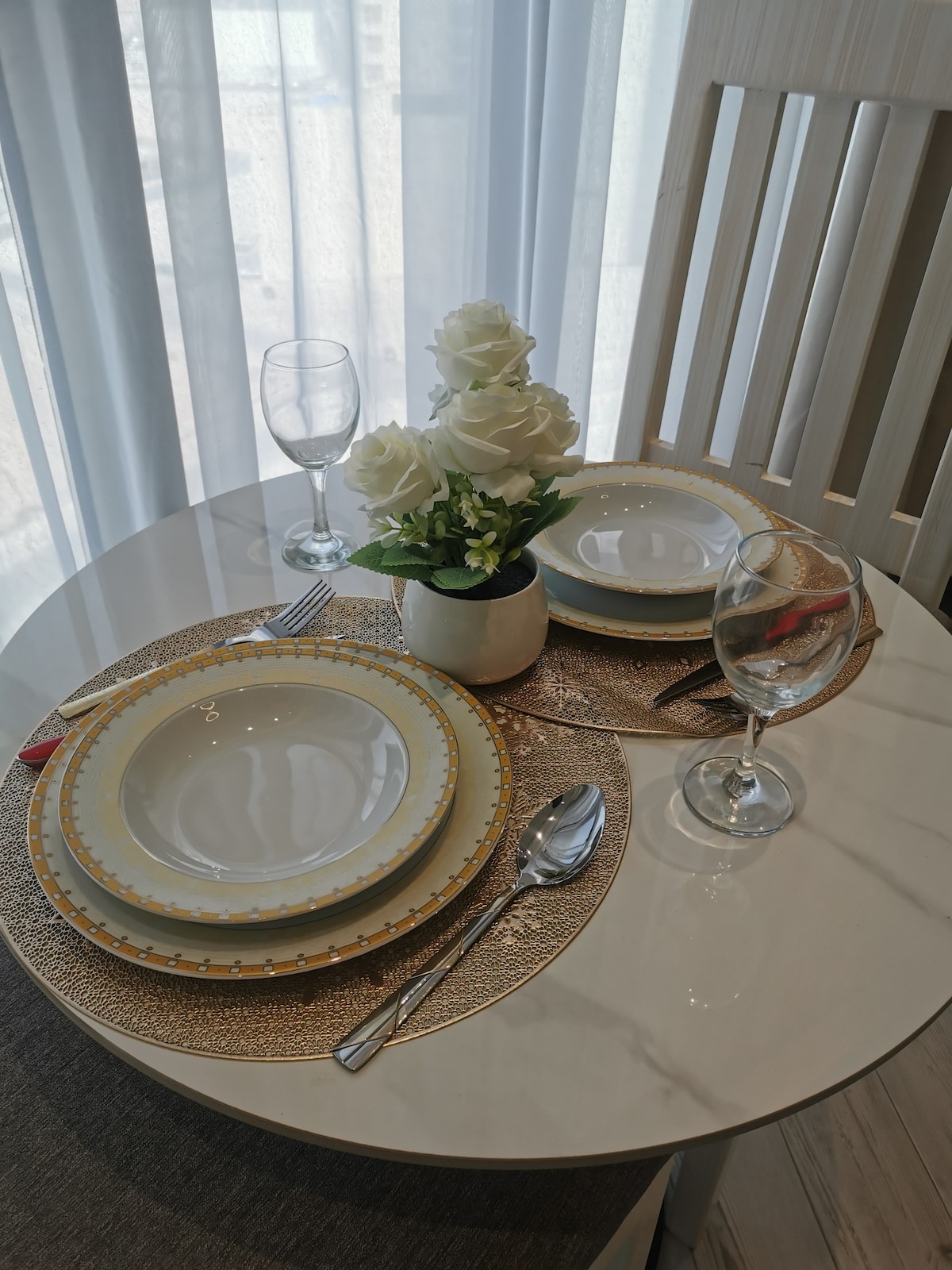 A dining table is set with elegant tableware, featuring round dishes with a gold rim, cutlery, and wine glasses. A small vase with artificial white roses adds a touch of decor. Natural light filters through sheer curtains, creating a bright atmosphere.