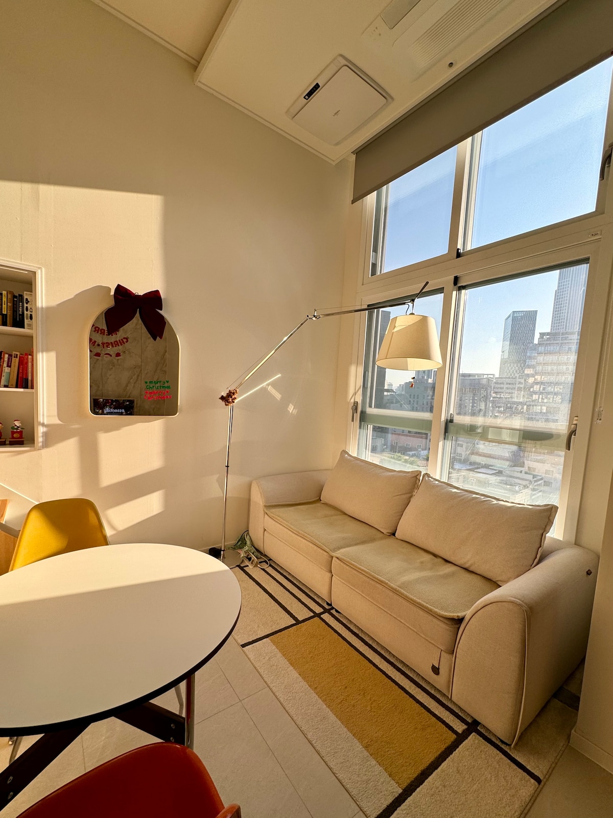 A cozy corner of the living space features a light-colored sofa positioned beside a window, allowing warm sunlight to fill the area. A round table sits nearby, accompanied by colorful chairs. A floor lamp adds soft illumination to the inviting atmosphere.