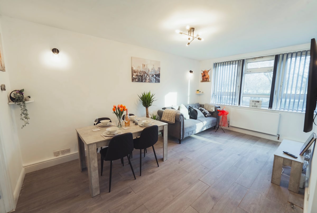 The living area includes a compact dining table set for four and a cozy sofa, thoughtfully arranged to encourage relaxation. Natural light filters through the vertical blinds, illuminating the space, while decorative elements like a potted plant and vibrant flowers add a touch of comfort.