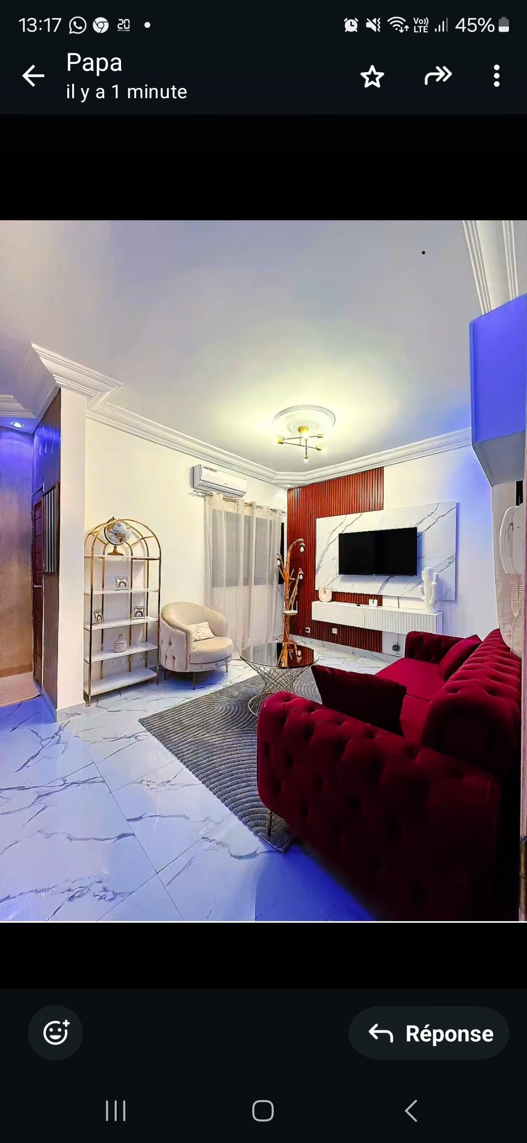 A modern living area features a comfortable red sofa and a stylish armchair. A sleek television is mounted on the wall opposite, complemented by decorative elements. Natural light filters through sheer curtains, while a striped rug adds texture to the marble floor.