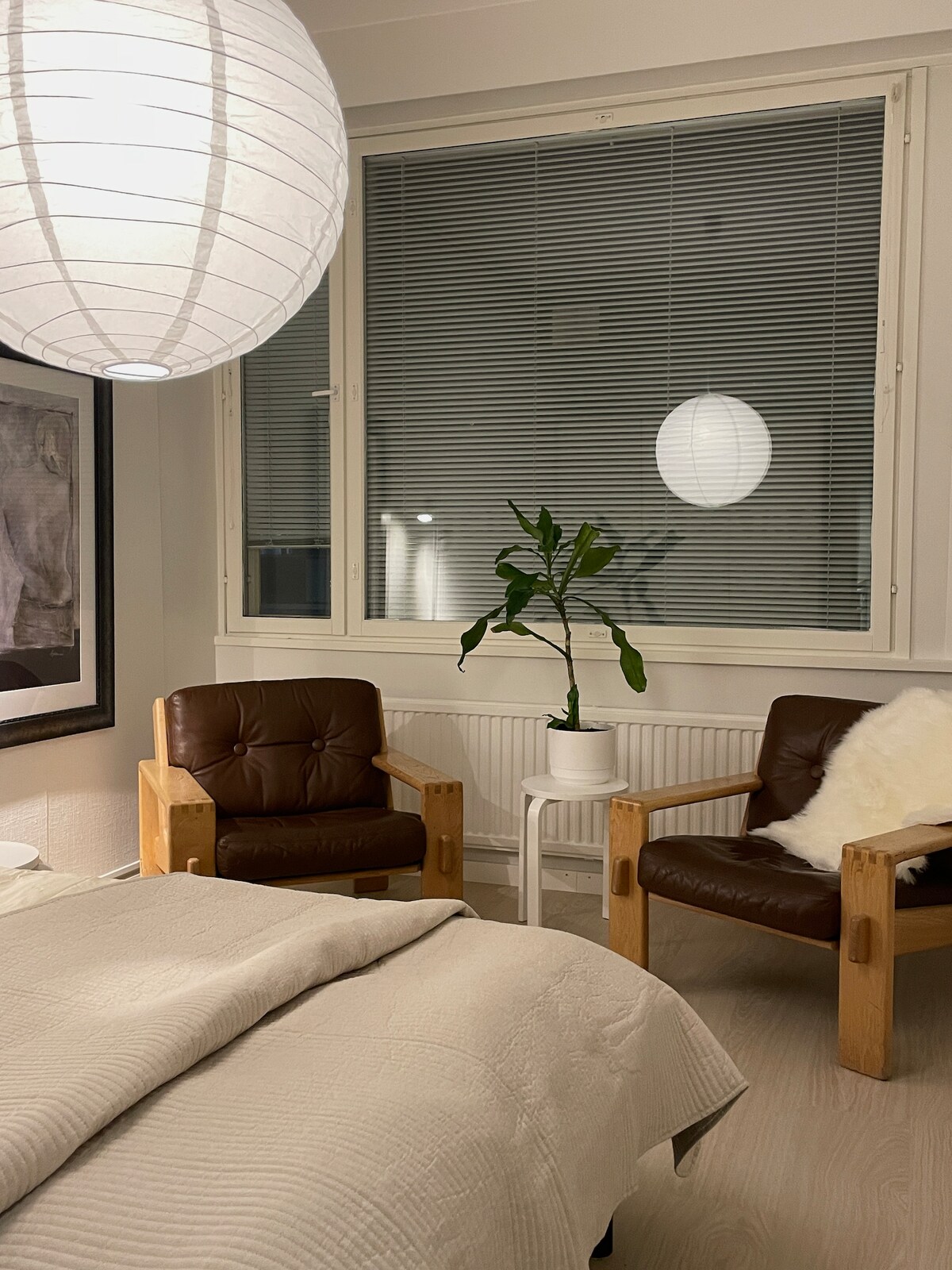 A light-filled bedroom features a spacious bed covered with a neutral-toned blanket. Two wooden armchairs with brown leather cushions are positioned near a small white table, accompanied by a potted plant. Natural light filters through the large window adorned with blinds.