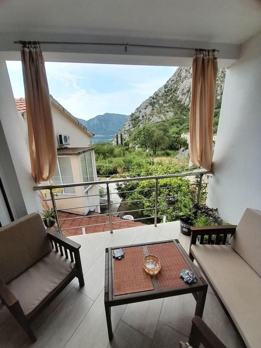 A private balcony features two wooden chairs and a small table. Lush greenery and mountainous terrain are visible through the glass railing, with soft, flowing curtains accentuating the space. The surrounding area is peaceful, showcasing elements of nature.