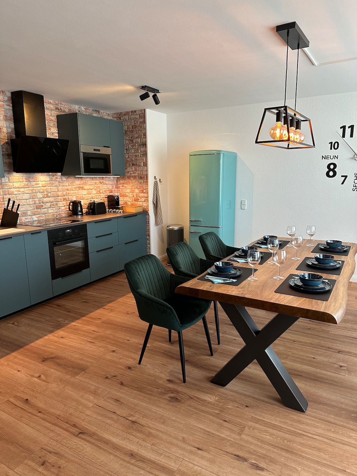 A modern kitchen is showcased with a striking blue refrigerator and sleek cabinetry. A wooden dining table with dark accents is set for six, featuring elegant dishware. Warm lighting highlights the textured brick wall, enhancing the room's contemporary feel.