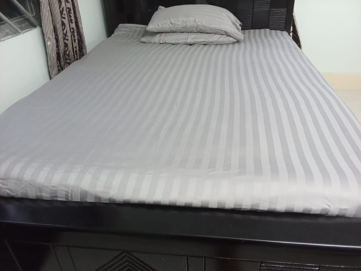 A spacious bed is presented with a striped gray coverlet and two matching pillows. Natural light is filtered through a nearby window, which is framed by patterned curtains. The bed is situated on a dark wooden frame that complements the room's decor.