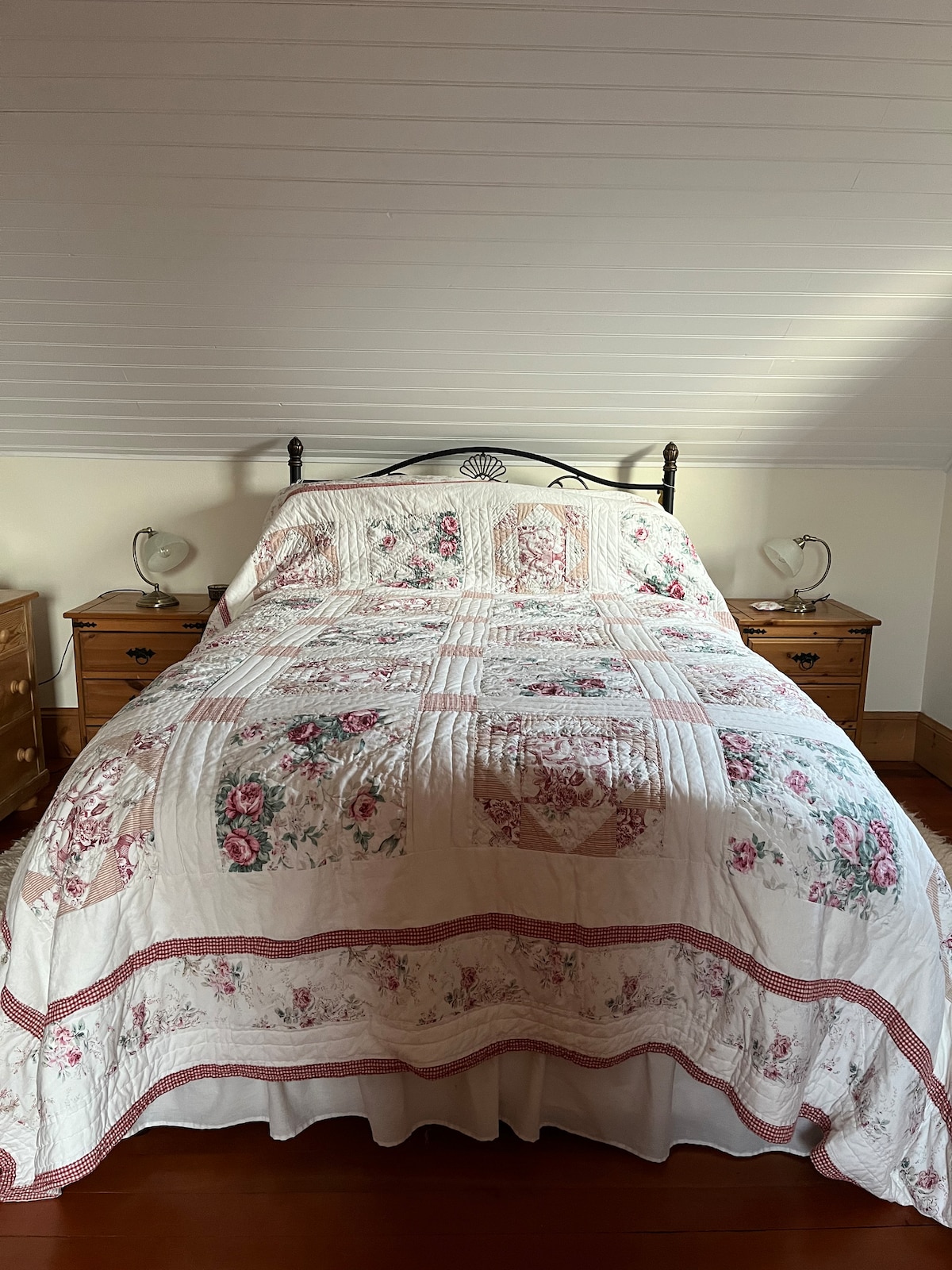 A spacious bed is covered with a floral-patterned quilt, complemented by two bedside tables with lamps on either side. Soft light enters from the window, illuminating the warm wood flooring and gently sloped ceiling.