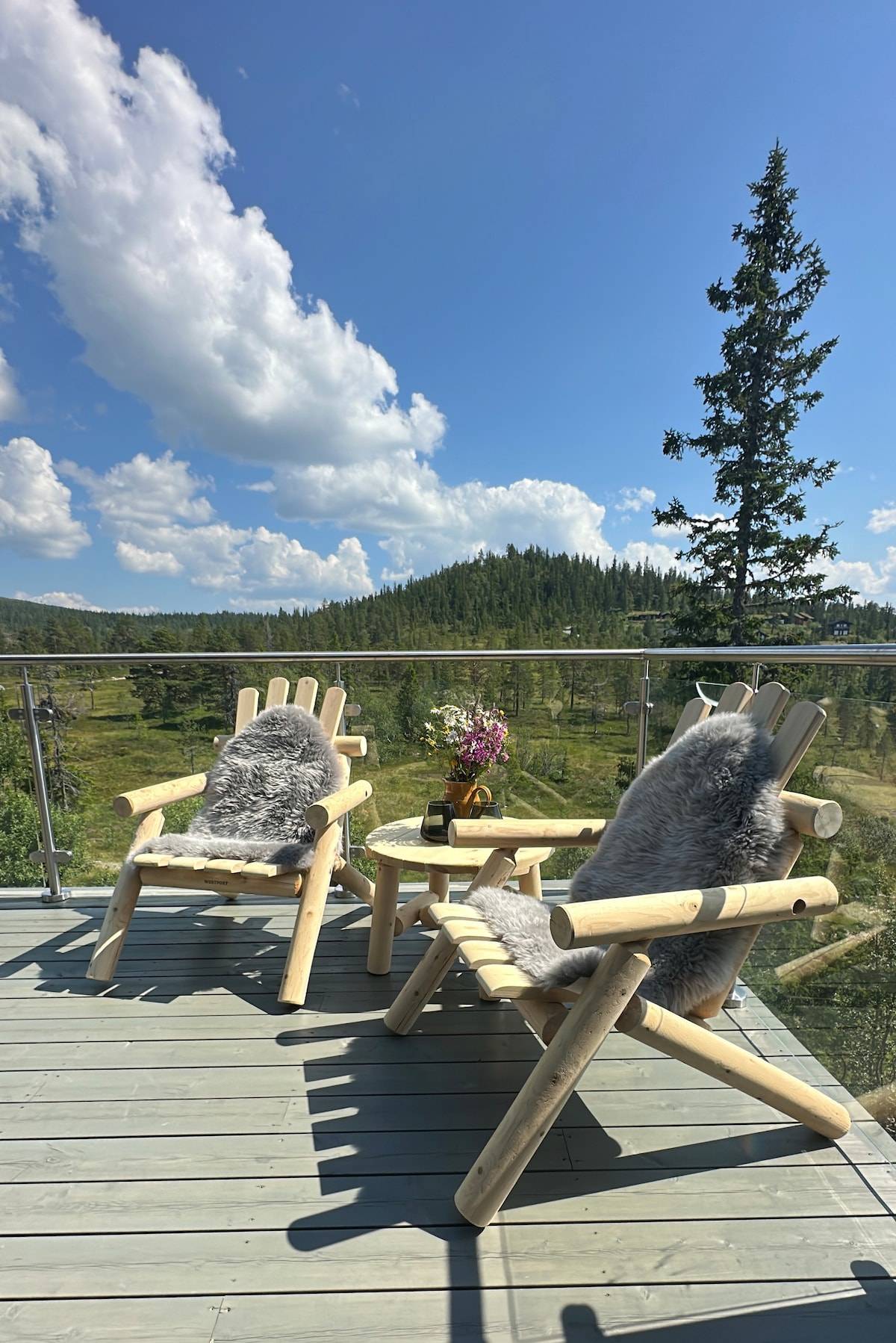 A sunlit terrace features two wooden chairs, each adorned with plush grey throws. A small round table, positioned between the chairs, holds a vase with flowers. The backdrop offers a scenic view of lush greenery and distant hills under a blue sky dotted with clouds.