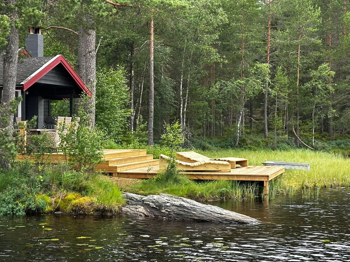 A wooden deck extends over the calm waters of a lake, surrounded by lush greenery and tall trees. A modest red-roofed cabin is visible nearby, with outdoor seating positioned to enjoy the tranquil setting.