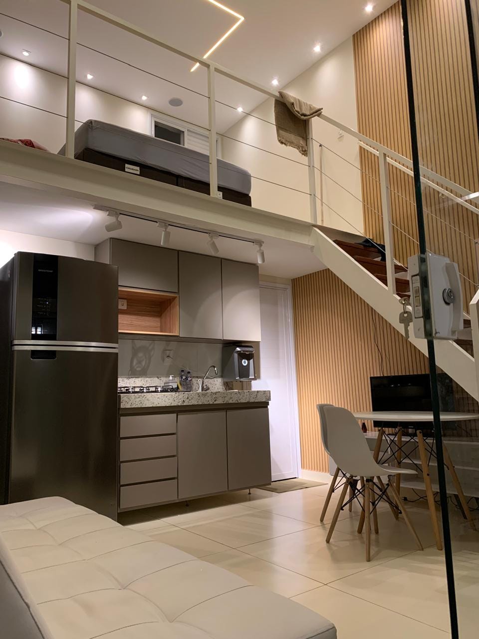 A sleek loft kitchen features modern appliances, including a refrigerator and cooktop, set against a stylish backsplash. A dining area with a table and chairs is included. The sleeping area is visible on the upper level, with a sectional sofa placed below. Ambient lighting highlights the space.