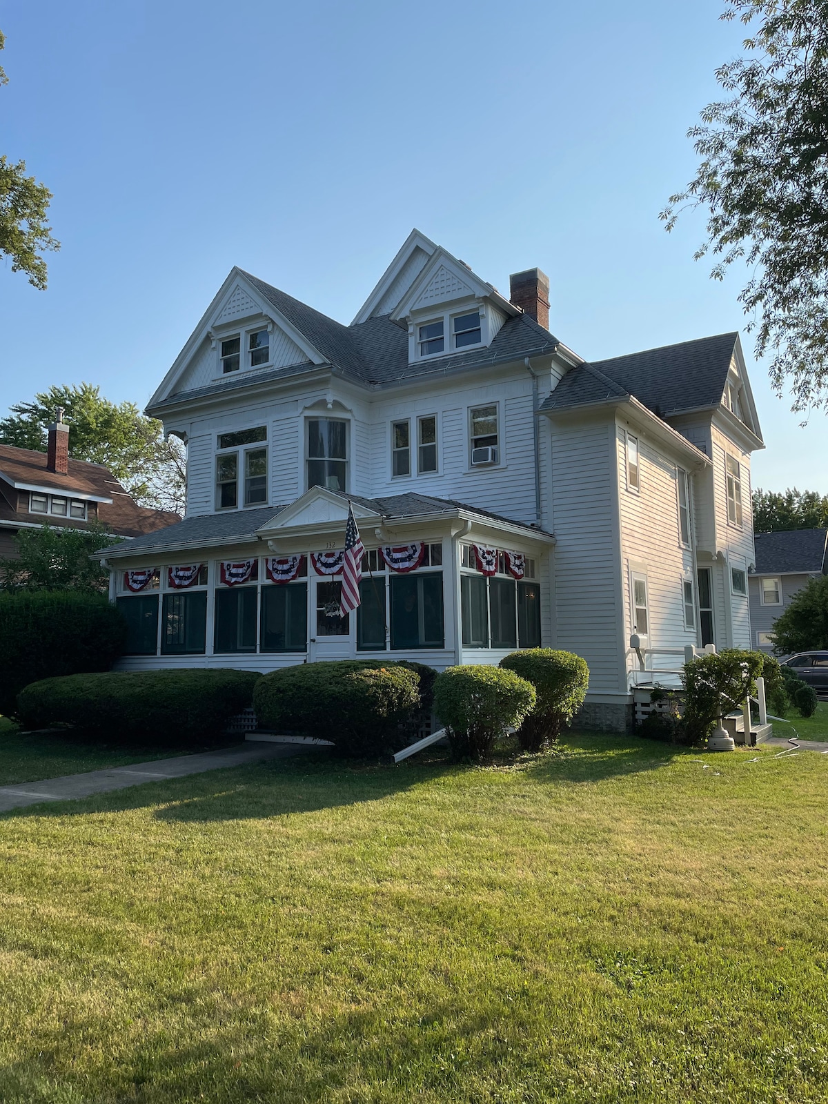 The exterior of a charming three-story house is shown, featuring a traditional design with a steeply pitched roof and decorative gables. A porch is visible, bordered by a manicured yard. American flags and bunting adorn the front, enhancing the welcoming appearance.