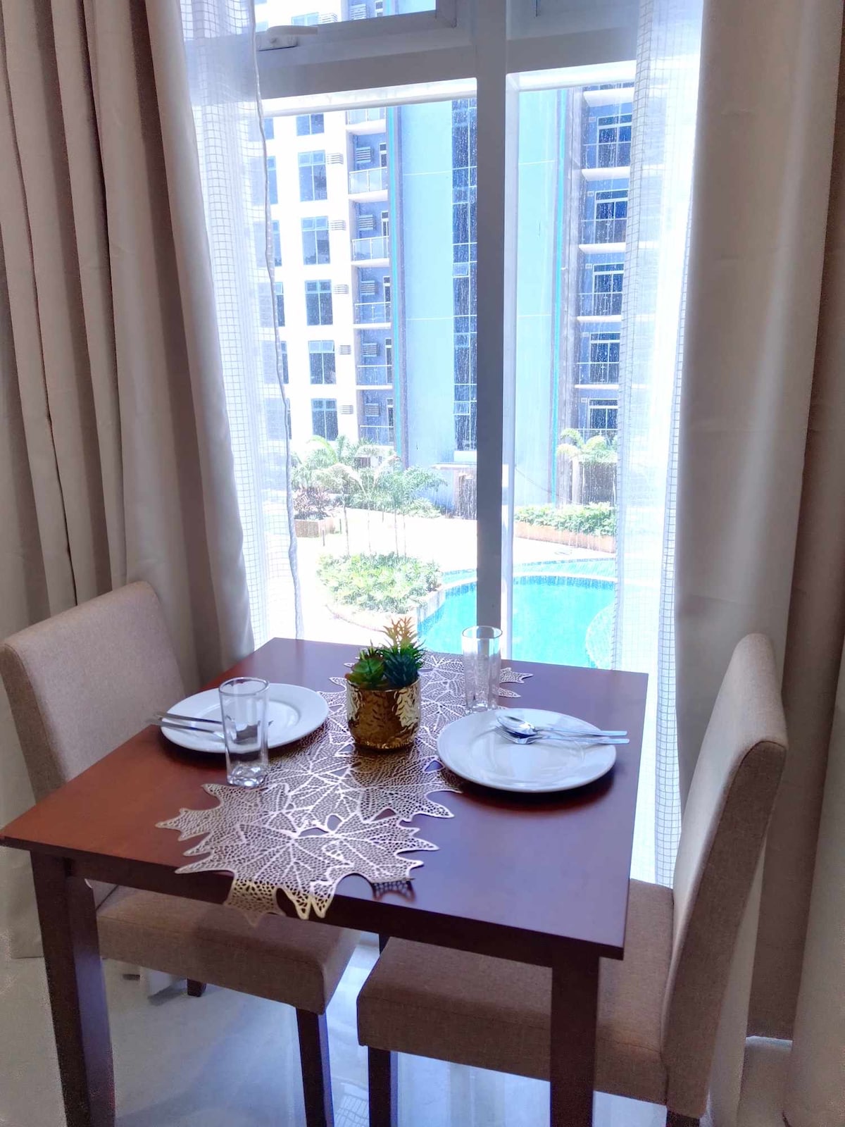 A dining area is set for two, featuring a dark wood table and beige chairs. A small decorative centerpiece is placed on the table. Large windows allow natural light to fill the space while offering a view of the pool and surrounding greenery.