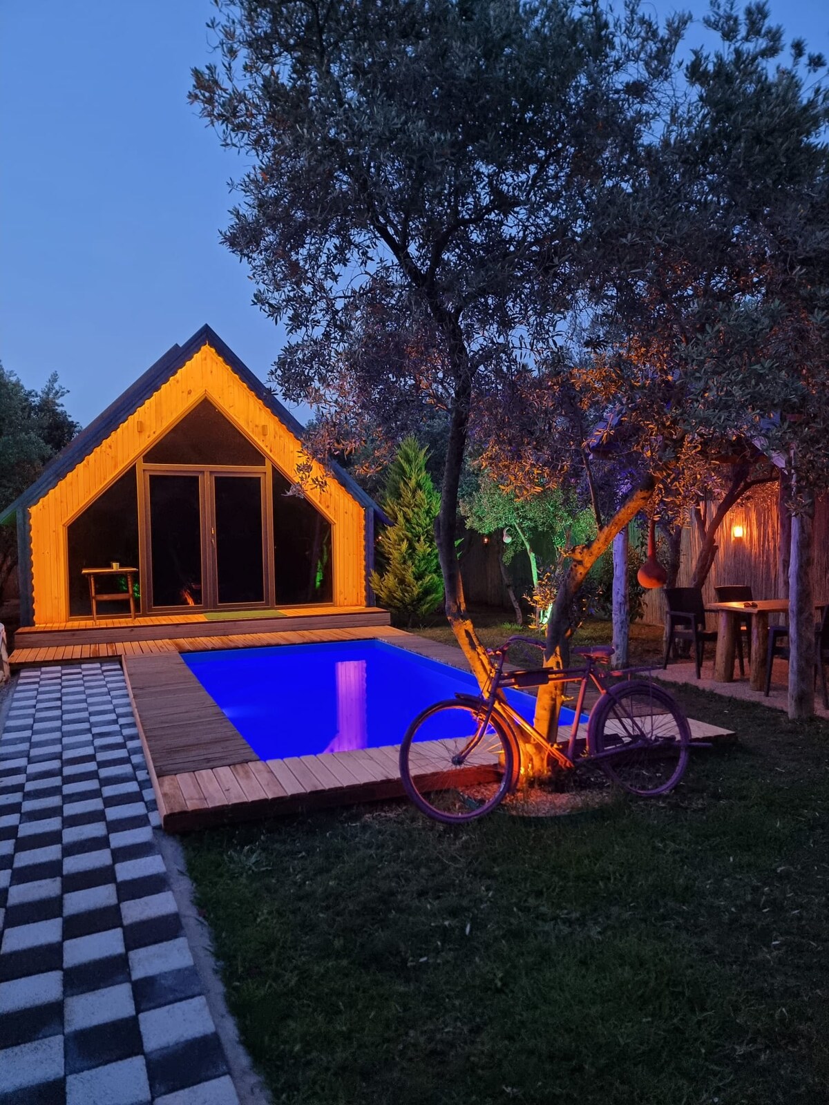 A wooden bungalow is set among olive trees, featuring a private pool with ambient lighting. A bicycle is placed near the poolside, and the area is surrounded by a well-maintained garden with natural greenery. The inviting outdoor space highlights the tranquil atmosphere.
