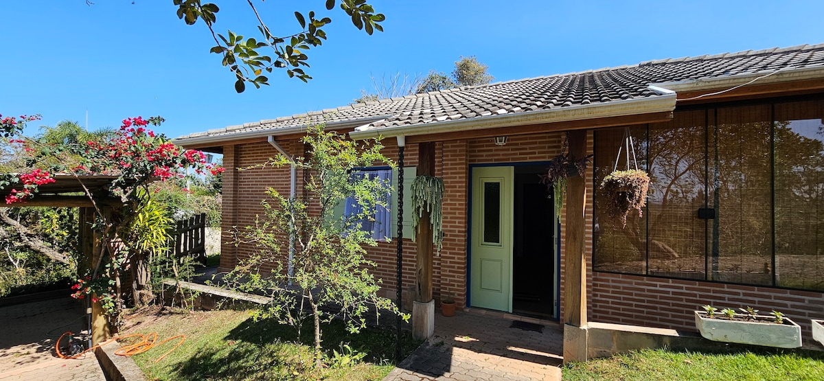 The exterior of a single-story brick house is showcased, featuring a green front door framed by hanging plants. A well-maintained garden with shrubs and flowering plants is visible, alongside a covered entryway. A clear blue sky provides a bright backdrop.