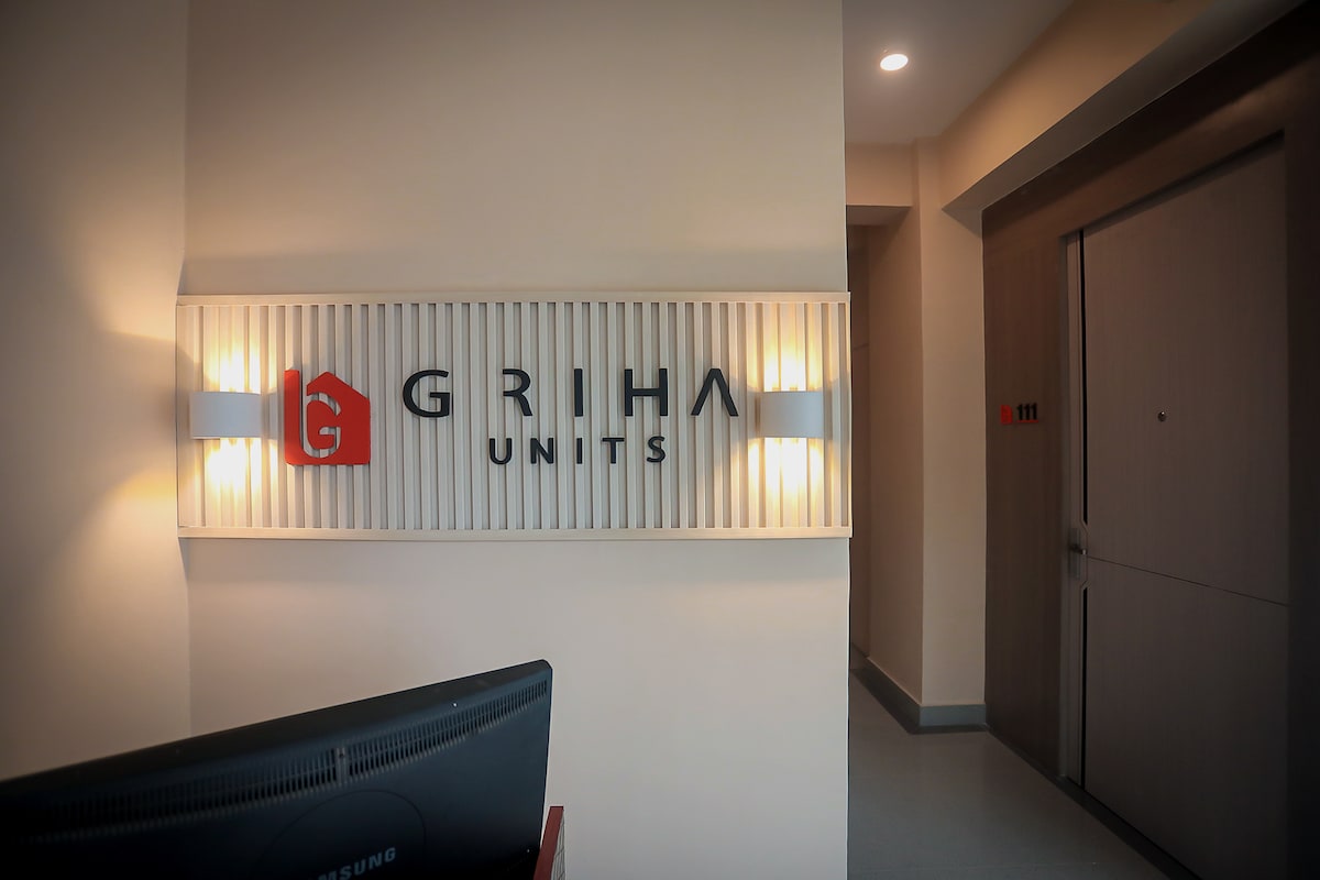 An illuminated sign displaying the name 'Griha Units' is mounted on the wall, complemented by soft ambient lighting. A reception area is visible, with a sleek desk positioned nearby and an elevator door shown in the background.