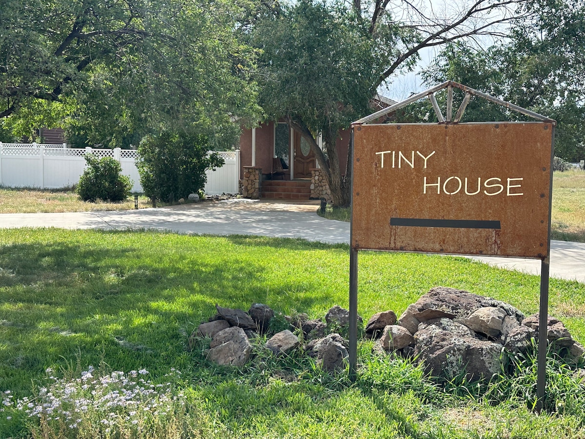 A rustic sign reading 'Tiny House' stands prominently in the foreground. A well-manicured lawn with green grass and boulders creates a natural setting. A charming house, partially obscured by trees, is visible in the background, complemented by a white fence.