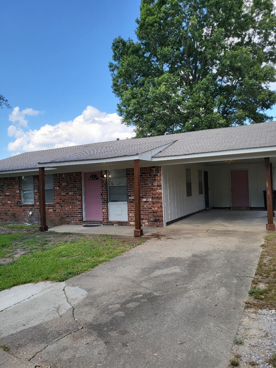 A modest single-story home with a brick exterior is showcased. The entrance features a pink door, complemented by a covered porch supported by wooden posts. A driveway leads to the home, flanked by a grassy area and a large tree providing shade.