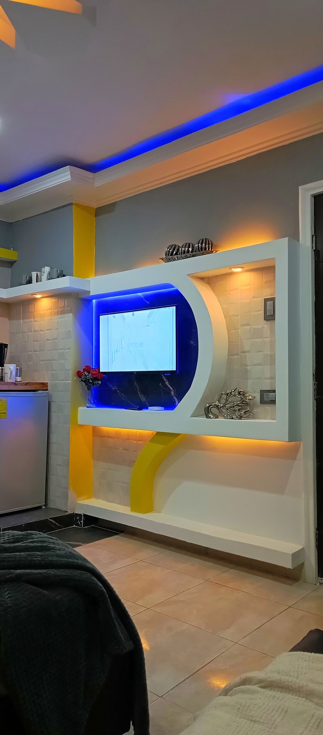 A compact kitchenette area features a refrigerator, microwave, and coffee maker, alongside a stylish white wall with integrated shelving. A mounted flat-screen TV is framed by a blue and yellow design, with soft lighting highlighting the surrounding elements.