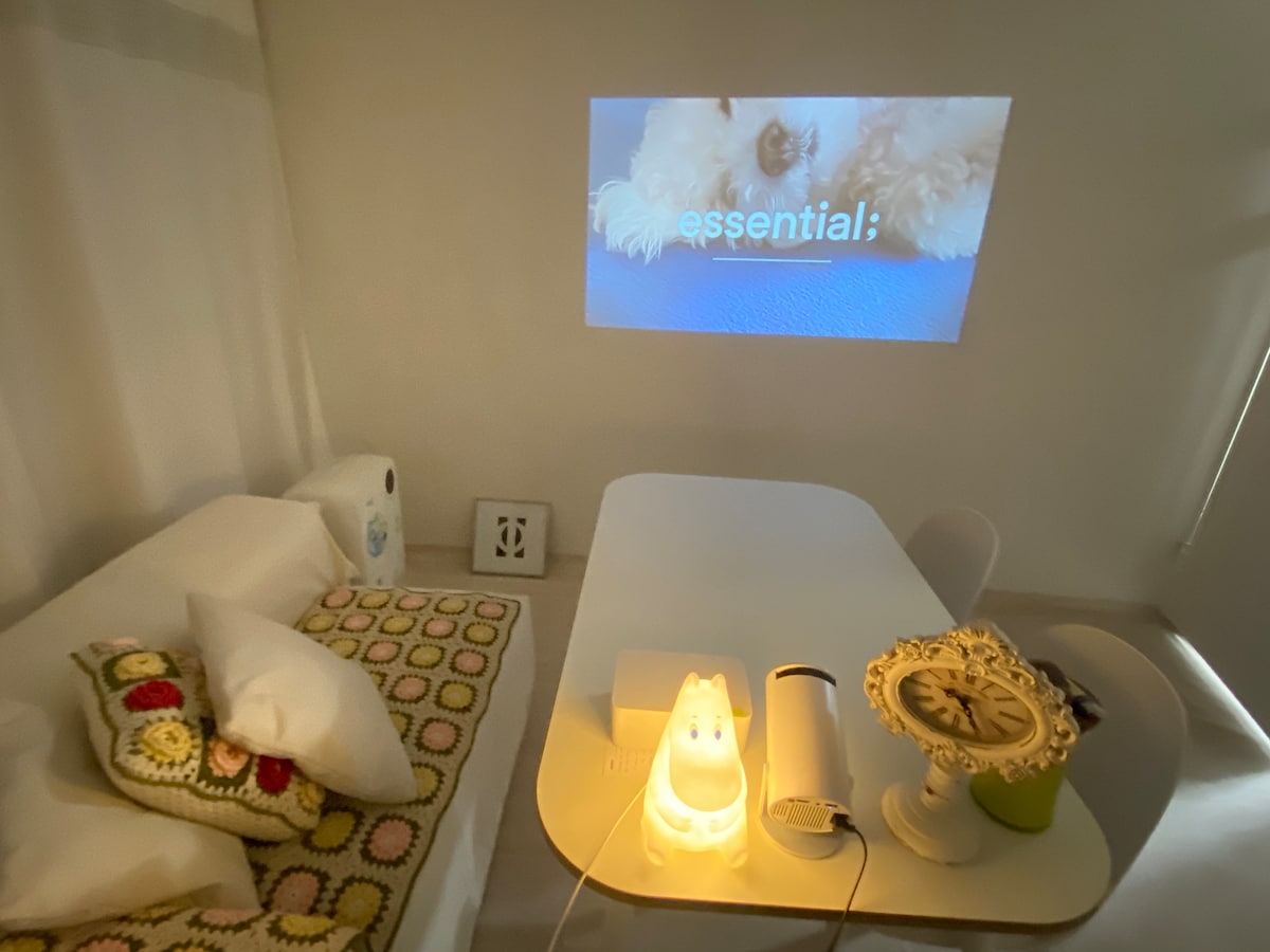 A cozy living area features a projecting screen displaying content against a wall. A table with chairs is positioned alongside a soothing nightlight and a decorative clock. Throws and pillows add comfort, while a colorful blanket lies draped over a sofa.