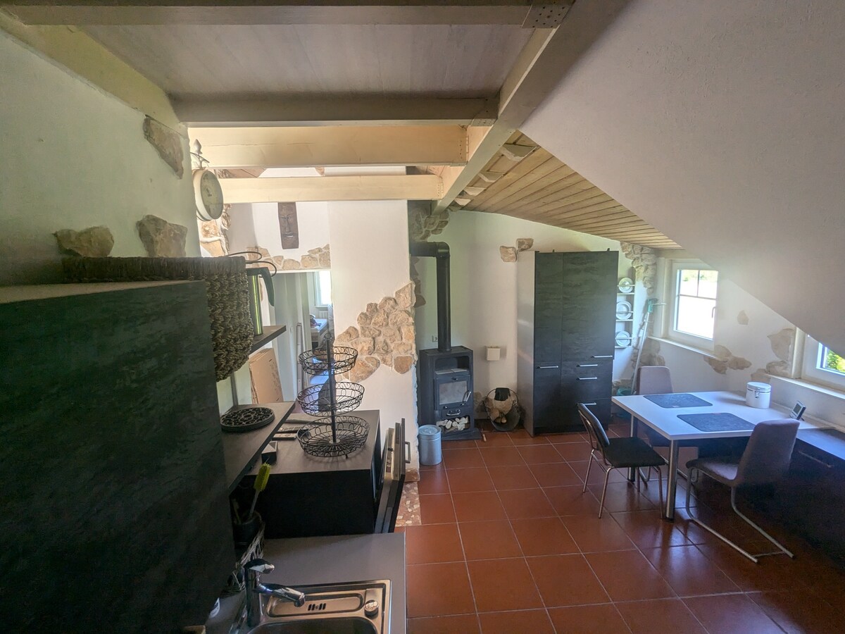 The kitchen area is highlighted by natural stone walls and wooden beams. A modern sink and stove are situated next to an inviting dining area with a table and chairs. Natural light fills the space through a window, enhancing the ambiance.