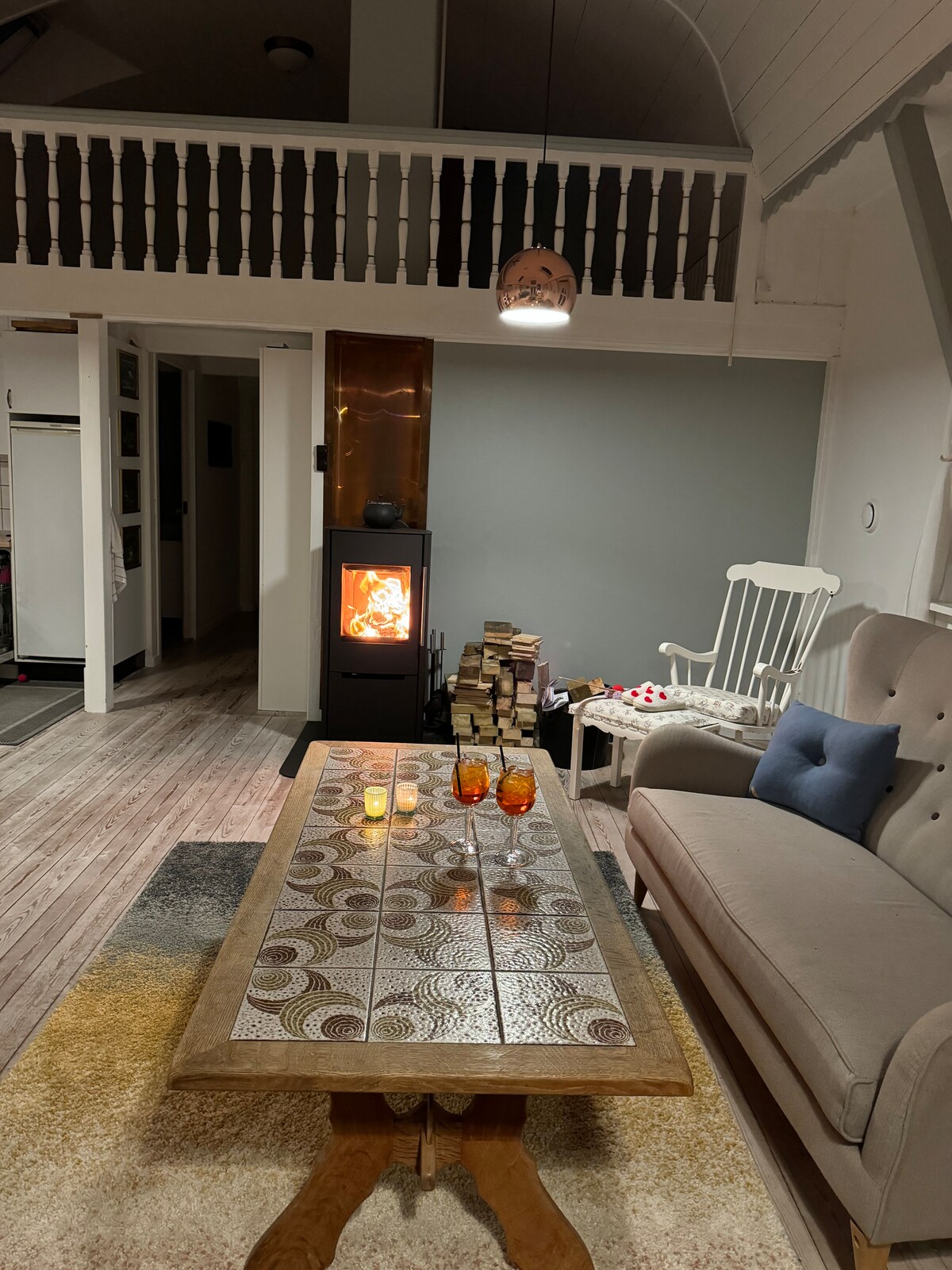A cozy living area features a soft couch accompanied by a styled table displaying drinks and candles. A modern wood stove provides warmth, while the walls are adorned with subtle decorations. Light fixtures enhance the inviting ambiance, and wooden flooring complements the space.