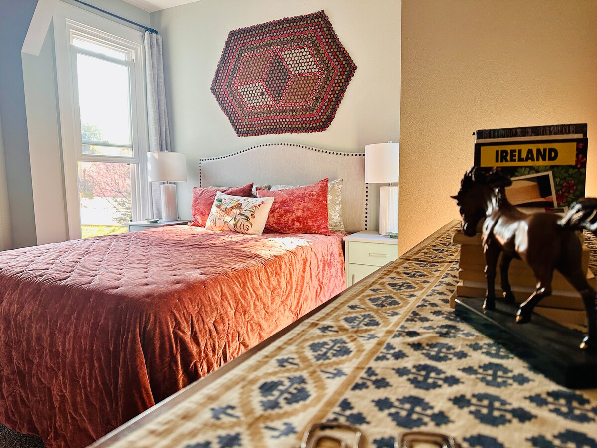 A cozy bedroom is presented, featuring a large bed dressed in a textured red coverlet. A colorful wall hanging adds visual interest above the headboard. Natural light filters through a window, illuminating a small decorative horse figurine and travel books on a bedside table.