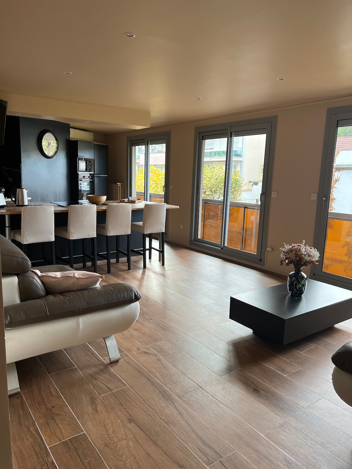 An open-plan living area is presented, featuring large windows that allow natural light to fill the space. A modern kitchen with bar seating is visible, complemented by a dining area. Light wood flooring enhances the overall ambiance, with a low coffee table centrally placed.