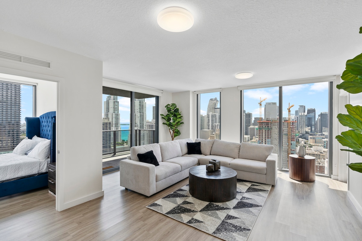 A spacious living area features large windows showcasing city and ocean views. A comfortable sectional sofa is arranged around a circular coffee table on a patterned rug. A green plant adds a touch of nature, while a bedroom area with a blue bed is visible in the background.