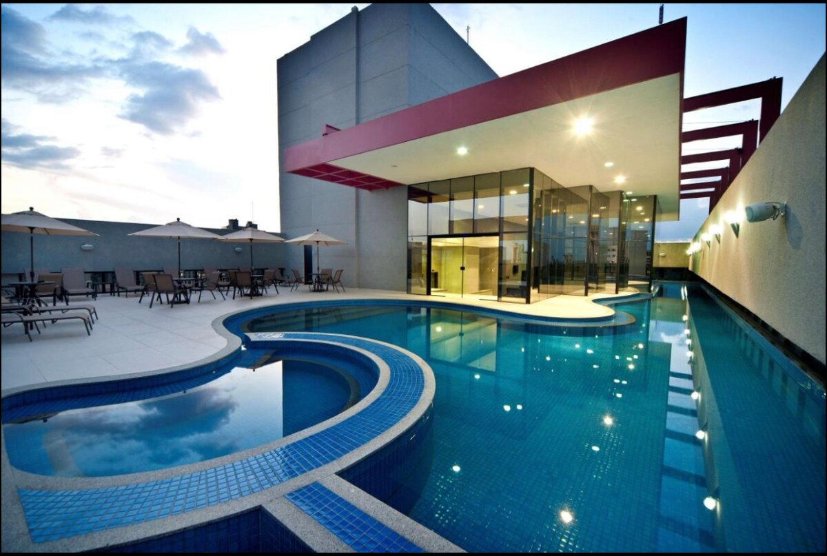 An outdoor pool area features a uniquely shaped pool with a blue mosaic border. Sun loungers with umbrellas are positioned around the pool. A modern building with large glass windows overlooks the area, reflecting the sky as evening approaches.