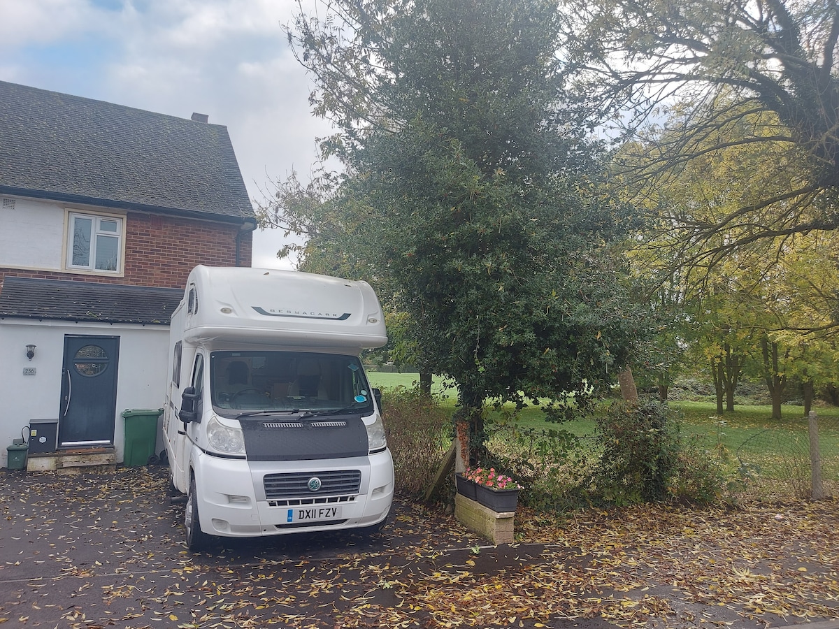 A motorhome is parked near a residence, surrounded by trees and foliage. The grassy area nearby adds a touch of greenery, while flower pots add color to the scene. The driveway is paved, with leaves scattered across the ground.