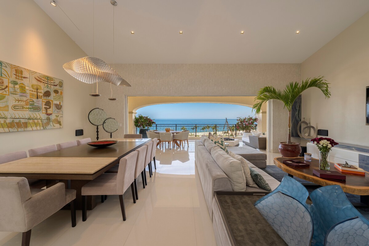 A spacious living and dining area showcases modern furnishings, including a long dining table and plush seating. Sunlight floods through large windows, illuminating the space and offering views of the ocean. Indoor plants add a touch of greenery, enhancing the inviting atmosphere.