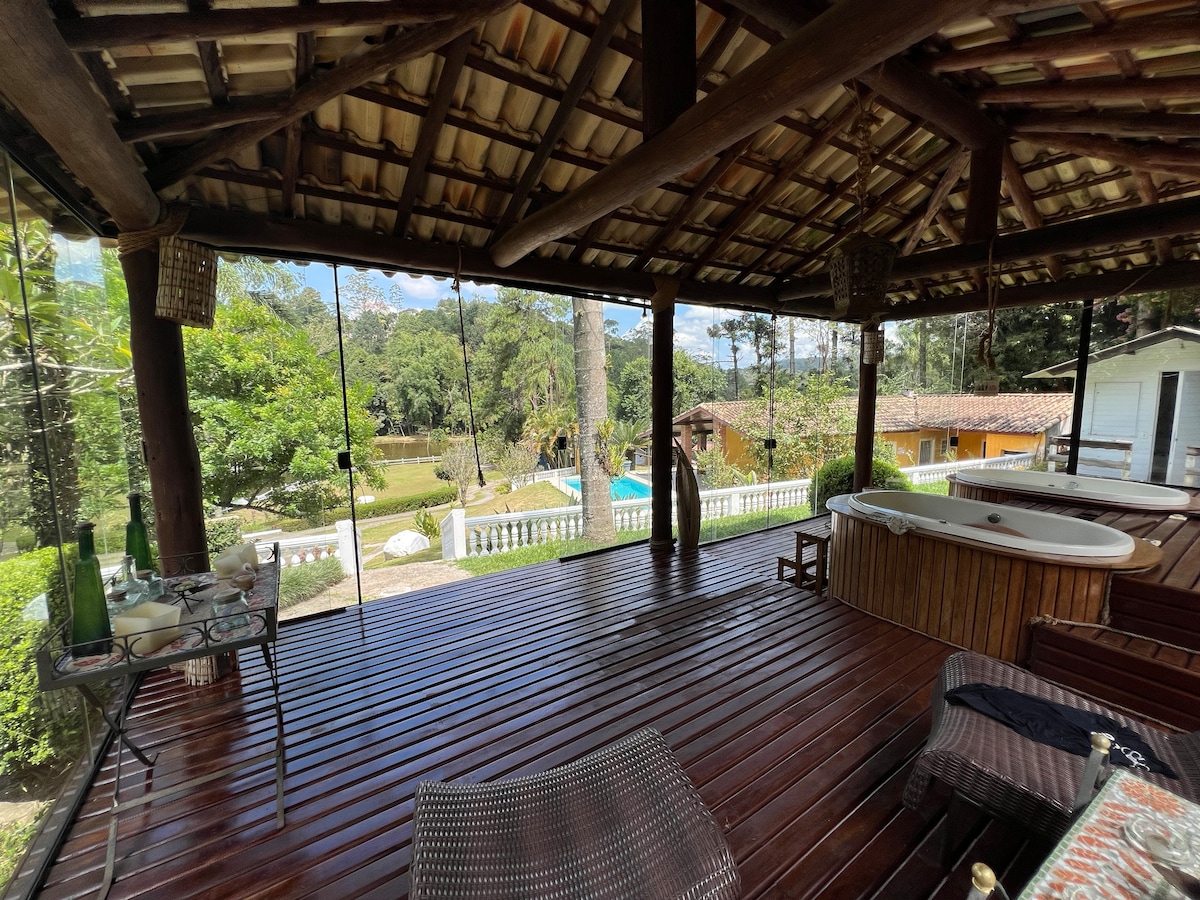 An open porch with a wooden floor features a jacuzzi and various seating options. Large glass windows provide views of the lush greenery and outdoor pool area. The rustic ceiling is supported by wooden beams, creating a spacious and inviting environment.